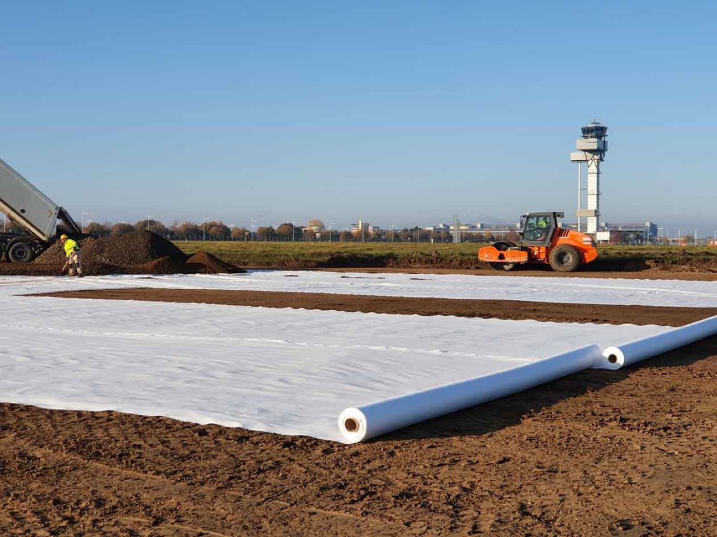 Un rouleau de géotextile non tissé blanc déroulé sur une plateforme préparée, avec un compacteur au travail à côté.