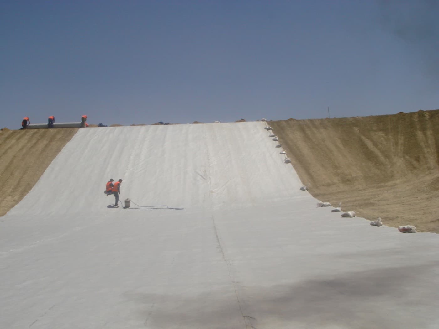 Géotextile posé sur un long talus de remblai, lesté en rive, avec deux ouvriers sur la nappe.